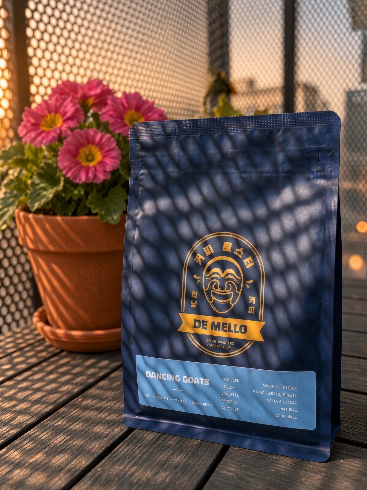 The Dancing Goats bag from De Mello Coffee Roasters, navy blue with a gold mask logo and a light-blue label, on a wooden table beside a pot of pink flowers, in low evening sun through a perforated metal screen.