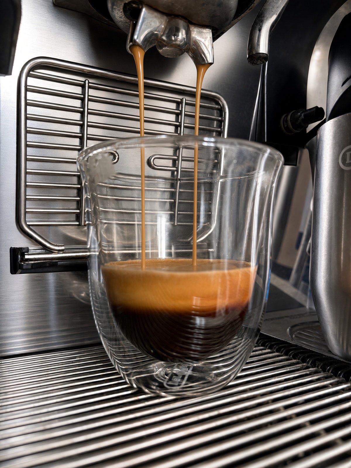 A double espresso pouring in two streams from the machine into a clear double-walled glass, under a thick golden crema, on the drip tray.