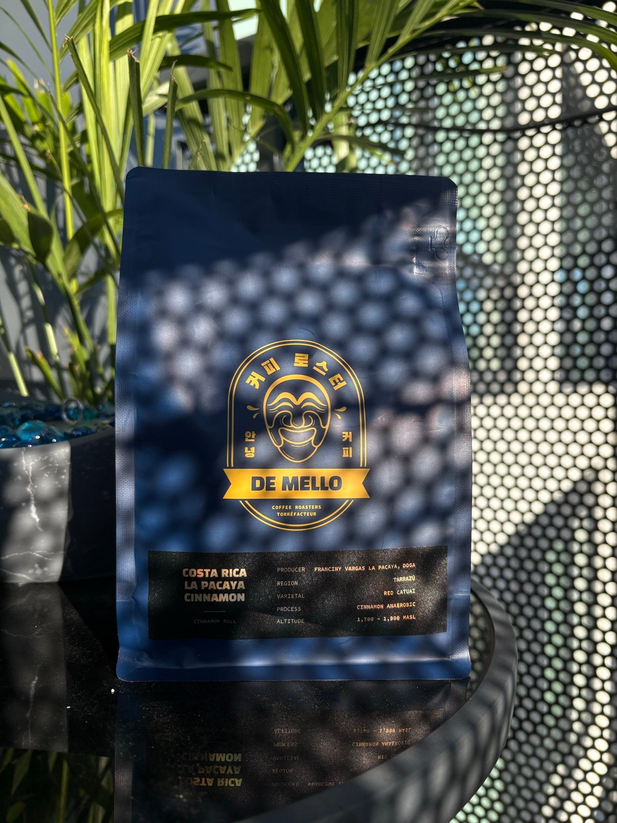 The La Pacaya Cinnamon bag from De Mello Coffee Roasters, navy with a gold label, on a black glass table beside a palm, dappled by the shadow of a perforated metal screen.