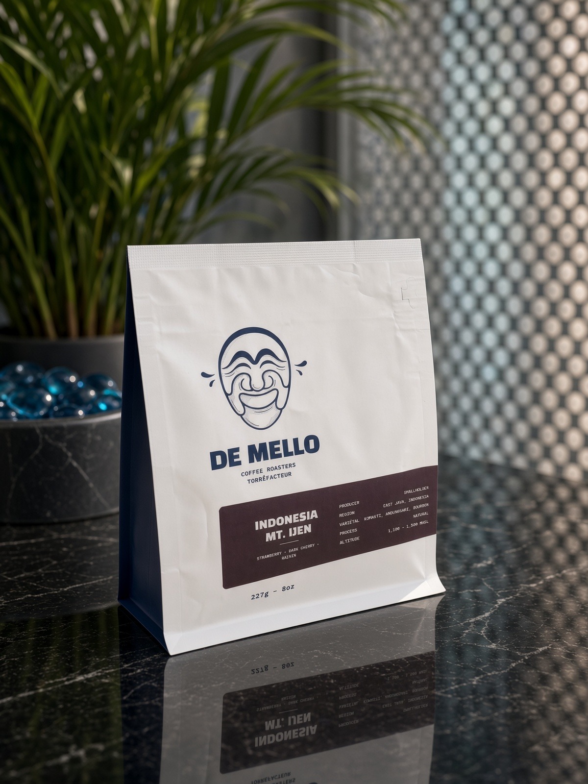 The Indonesia Mt. Ijen bag from De Mello Coffee Roasters, white with a maroon label, on a black marble counter beside a palm, in front of a perforated metal screen.